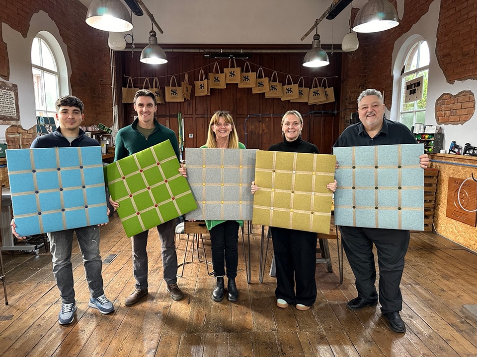 A group of colleagues standing together inside the chapel workshop studio, laughing and talking during a creative team-building activity, with wooden tables and stained-glass windows in the background.
